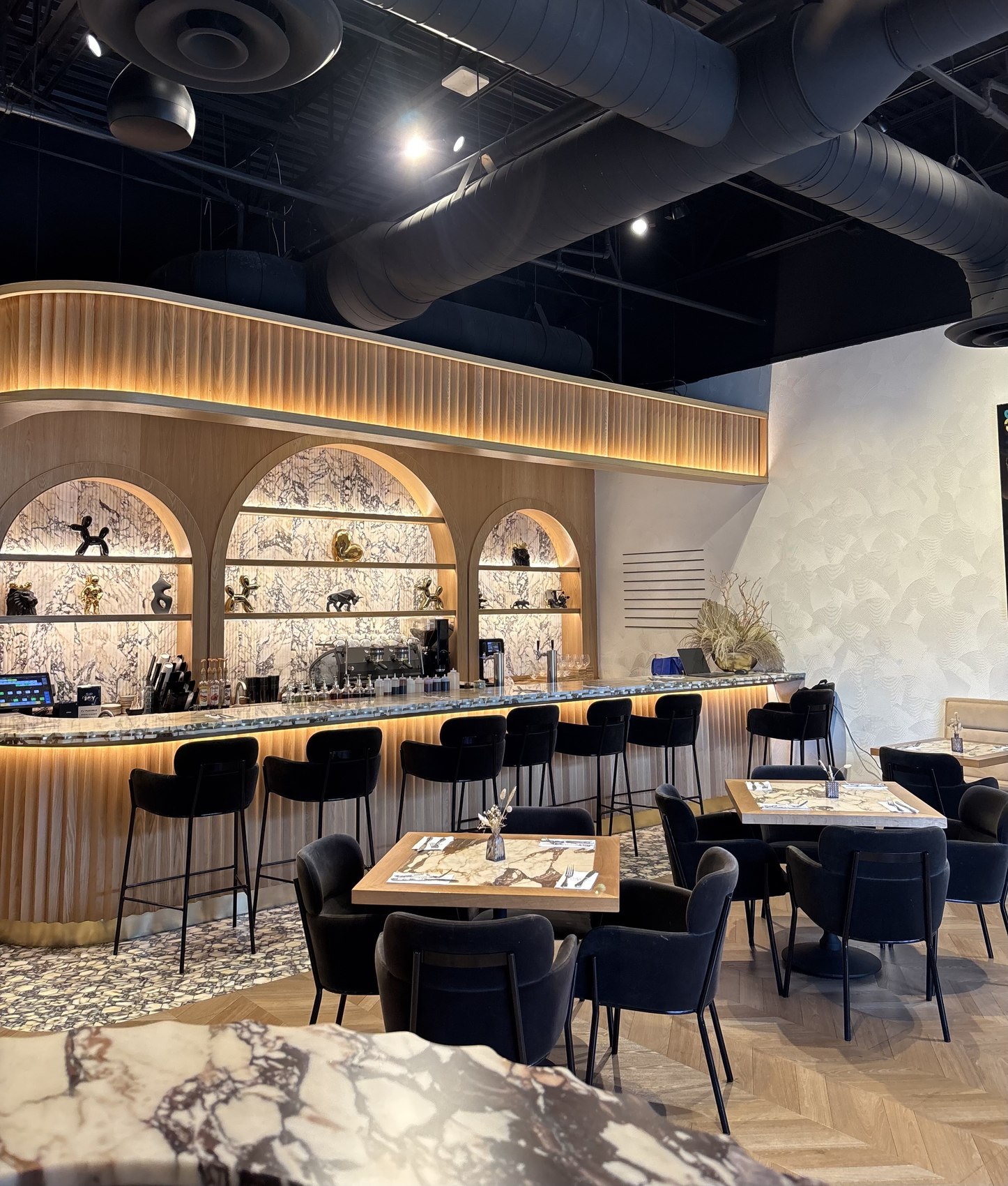 The curved coffee bar and bar stools at Crave Café, with marble-topped tables and the lit arched shelving behind
